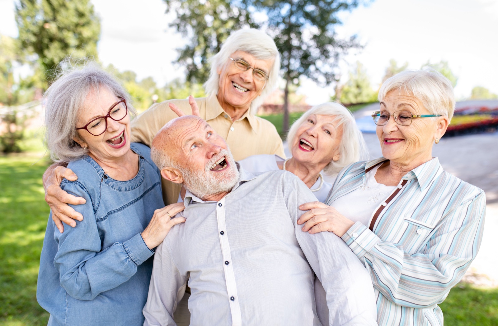 A group of 5 older friends standing outside in a park, making silly faces and having fun.