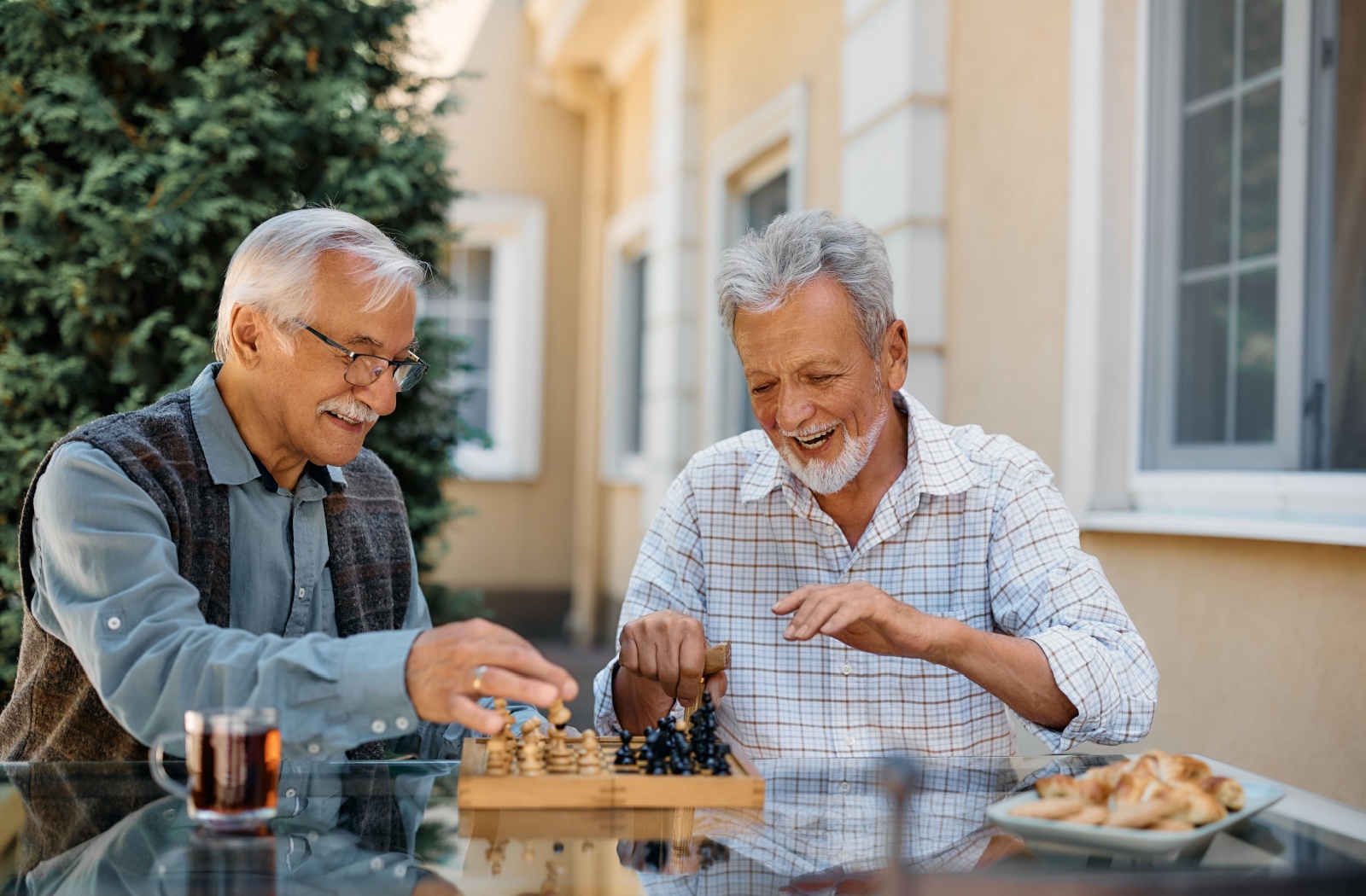 A pair of older friends sitting outside on a patio in senior living, laughing over a game of chess together.

