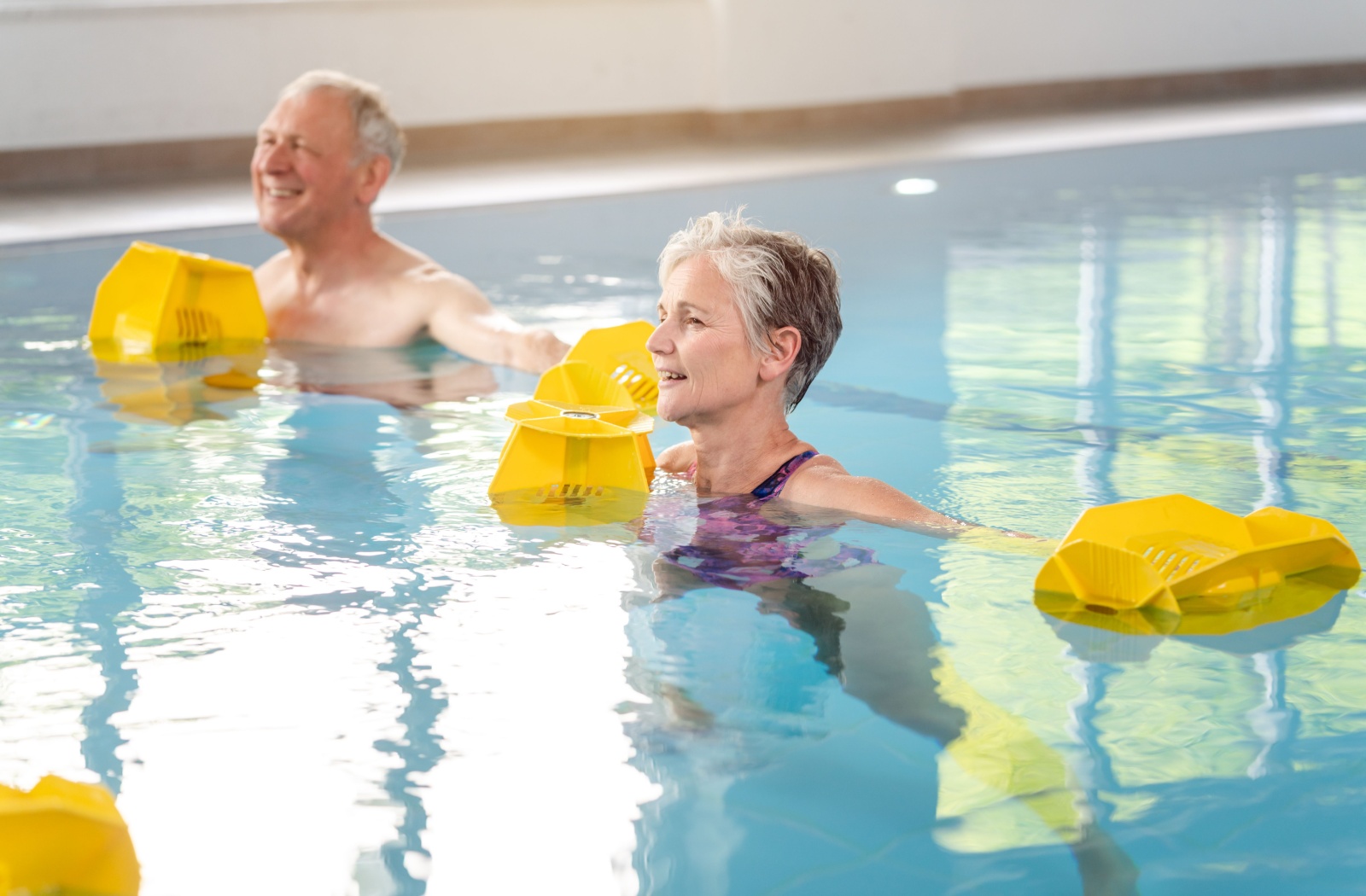 A pair of older adults with plastic floatation devices swimming in a pool during aquatic therapy.