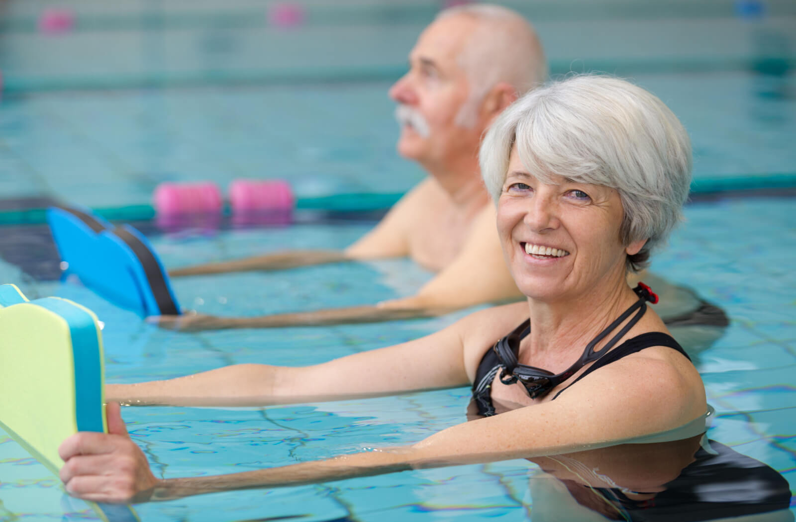 An older couple in senior living holding paddleboards in the pool, smiling during aquatic therapy.
