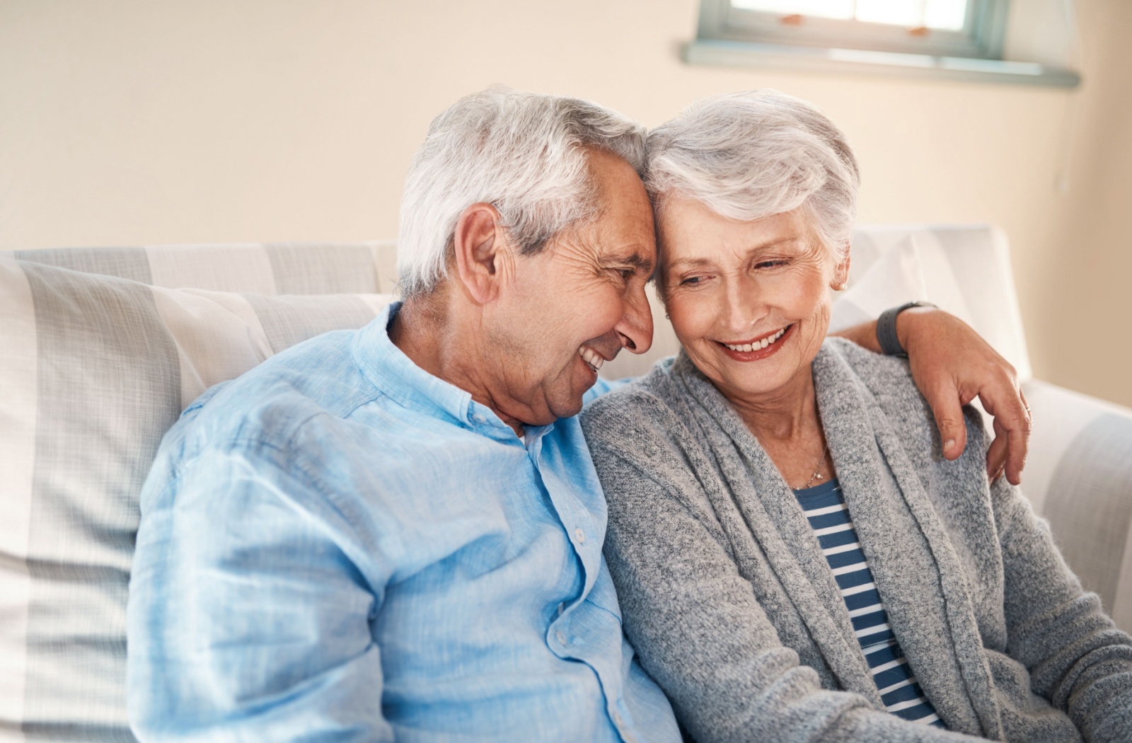 A senior couple cuddle together on the couch, smiling at each other, happy to have another day together in senior living