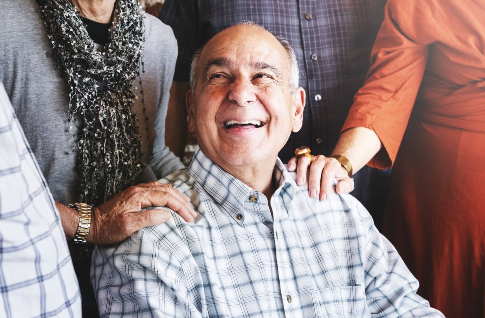 A seated senior smiles happily, surrounded by friends with their hands on their shoulders, representing community
