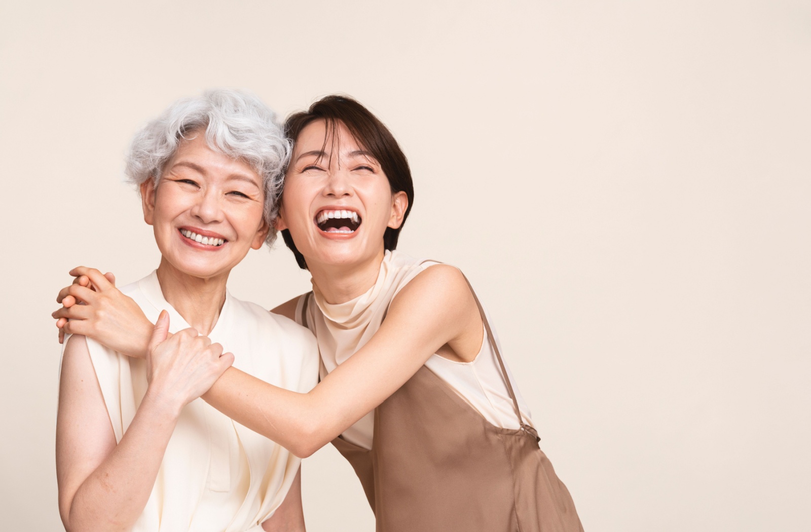 Against a beige backdrop, a smiling senior is embraced by their laughing, adult child, representing their close relationship
