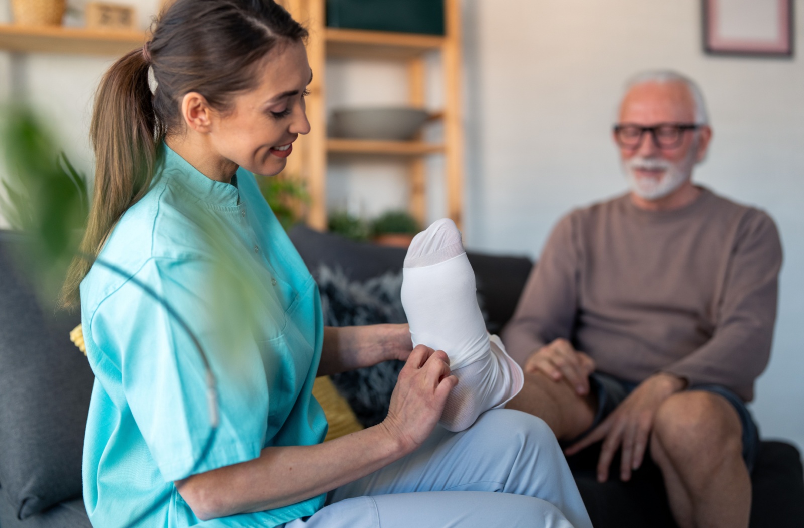 A smiling caregiver helps an older adult in assisted living try on a compression sock to help them deal with knee discomfort