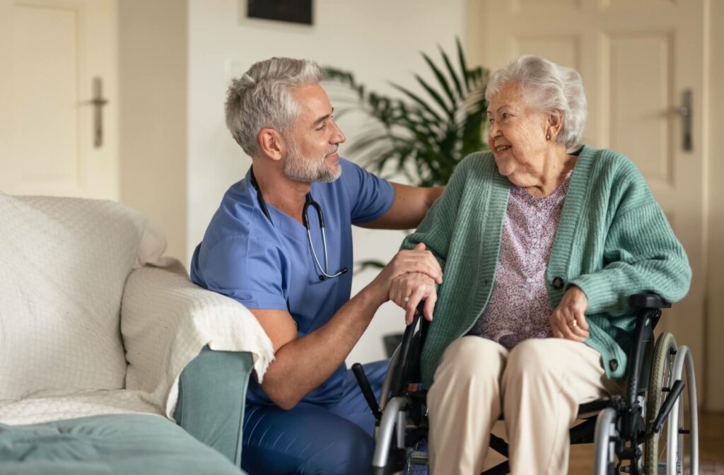 An older woman in a wheelchair and a male nurse crouching next to her and they smile at each other