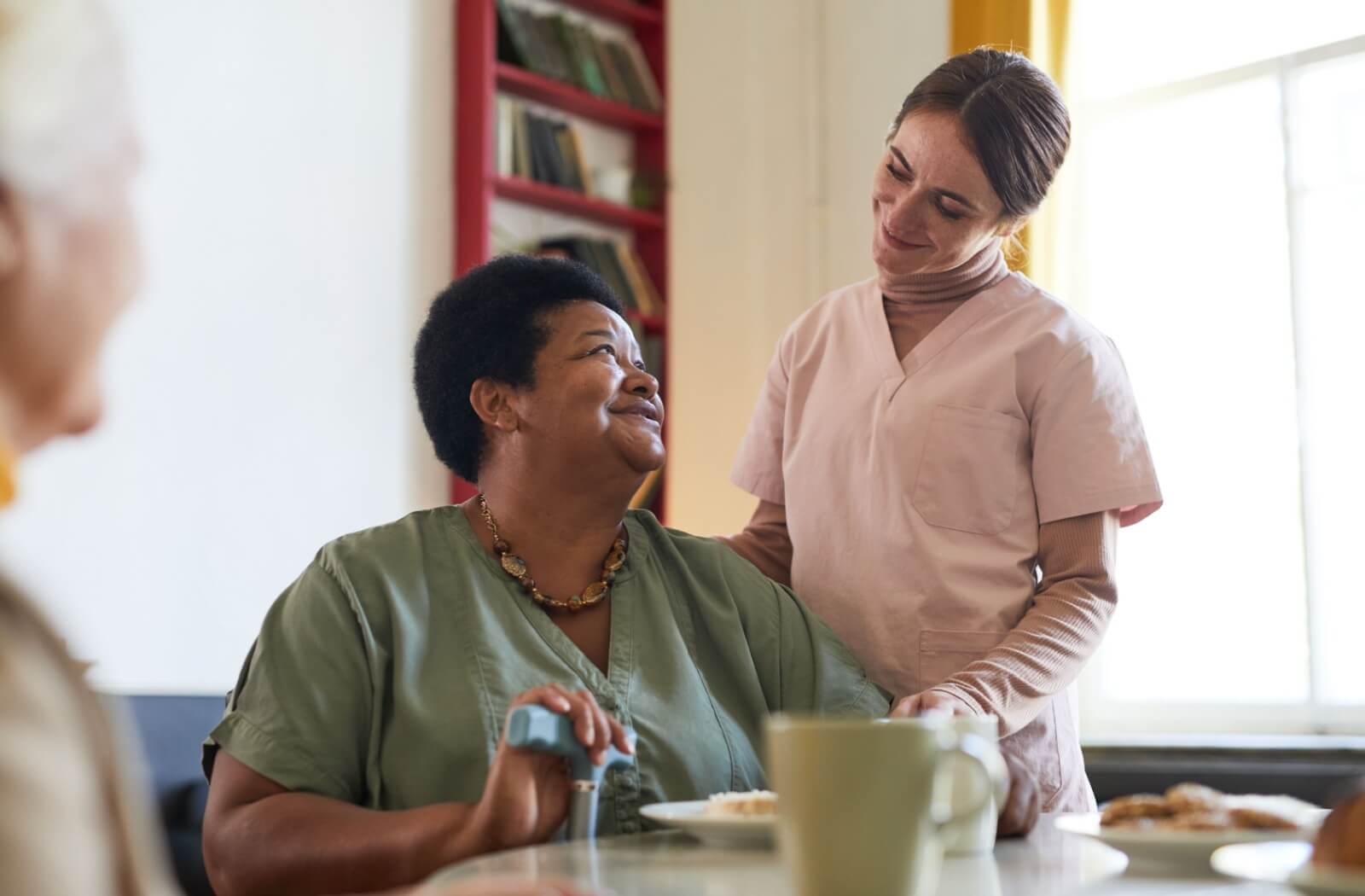 An older woman sitting at a dining table with a female nurse standing behind her and they smile at each other