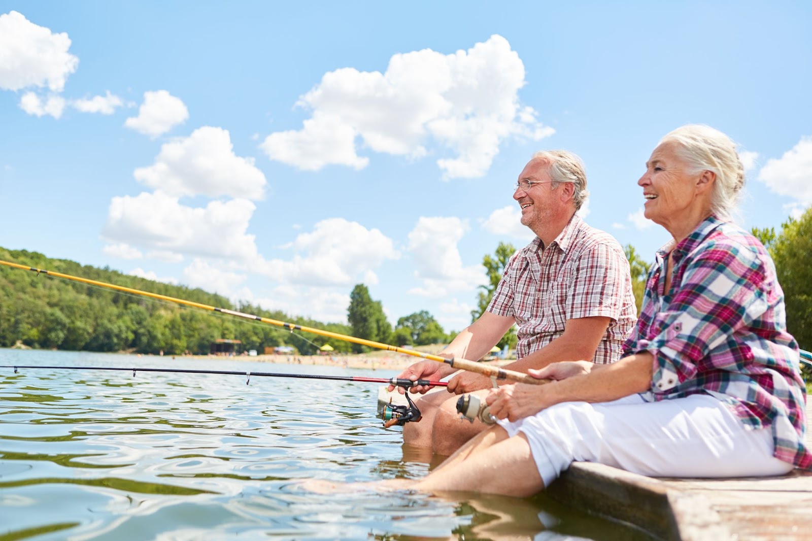 Two older adults sit on a platform on a sunny day with their legs on the water and fish.