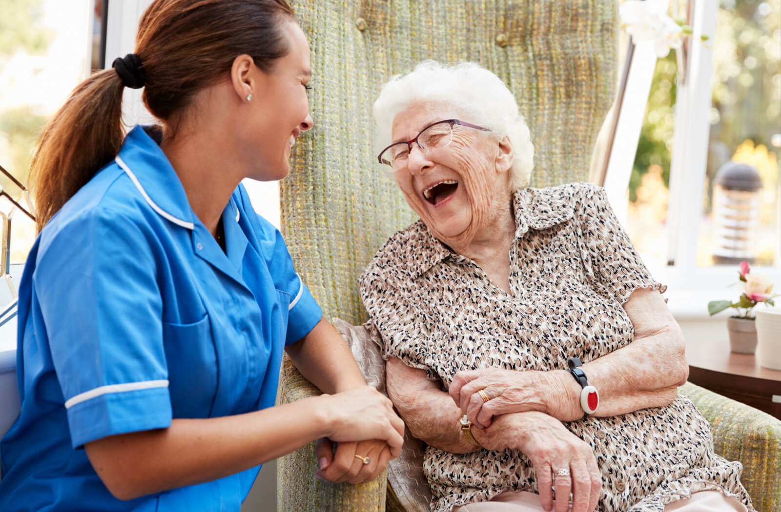 A caregiver kneeling beside an older adult with dementia in a chair in memory care, smiling and laughing together.