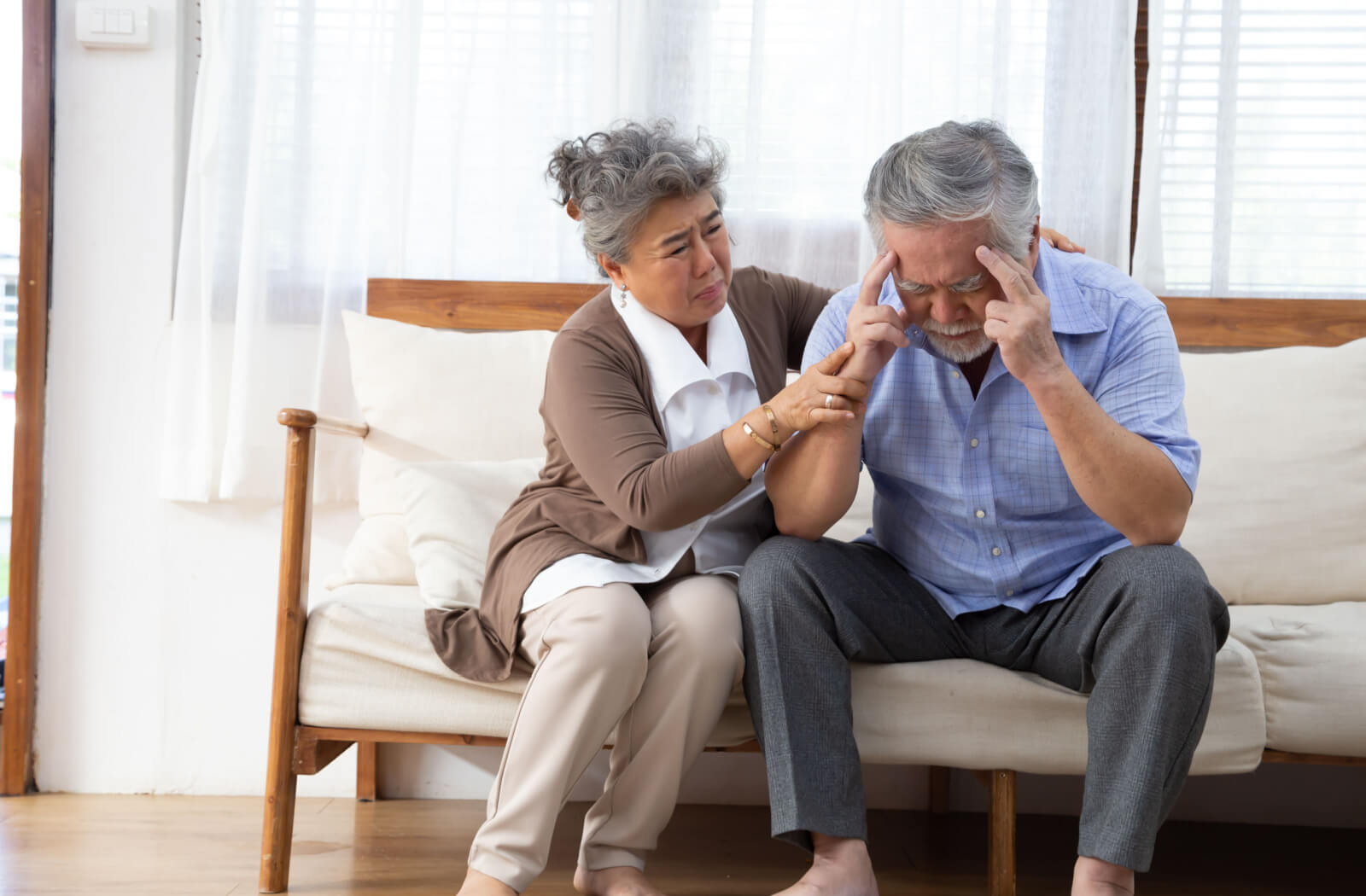 Seated together on a couch, a wife gently comforts her husband, who is visibly irritated and grappling with the challenges of recalling his memory.