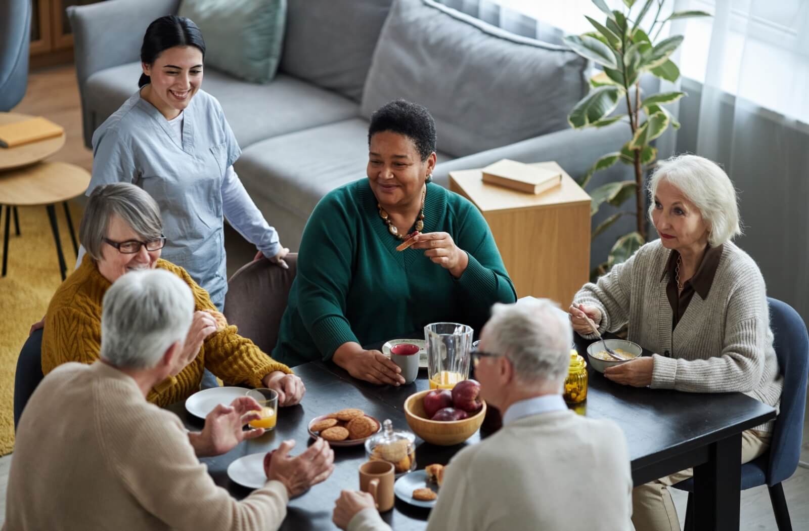 A group of older adults enjoying breakfast in respite care while a caregiver checks in on them.