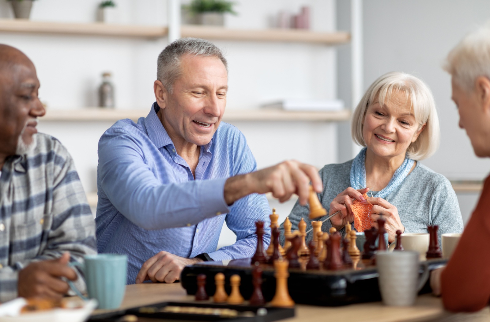 A group of older adults with limited mobility sitting around a table in senior living and laughing while playing chess.