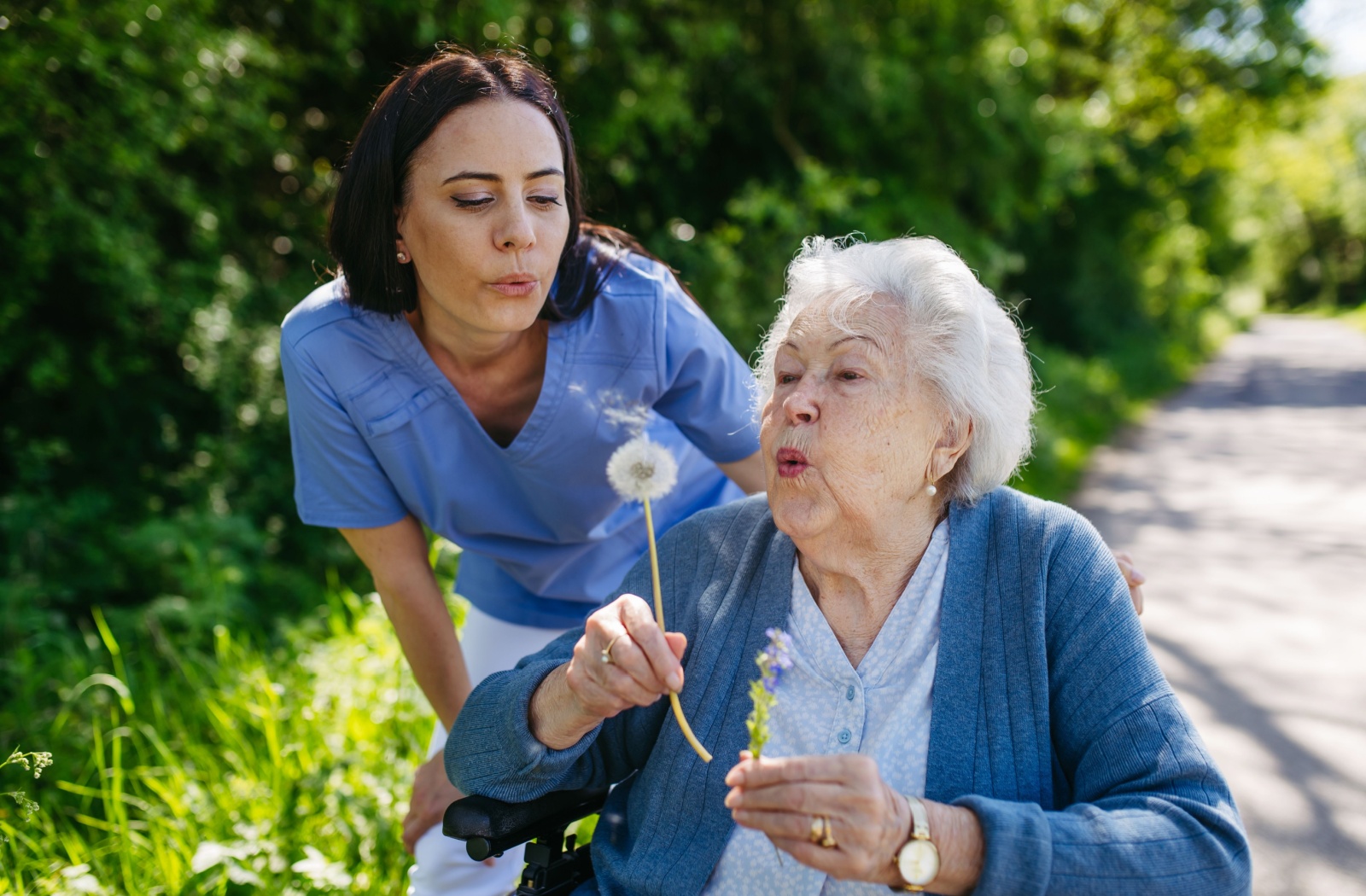 A caregiver and an older adults outdoors in a common garden, holding a dandelion and blowing the seeds away.
